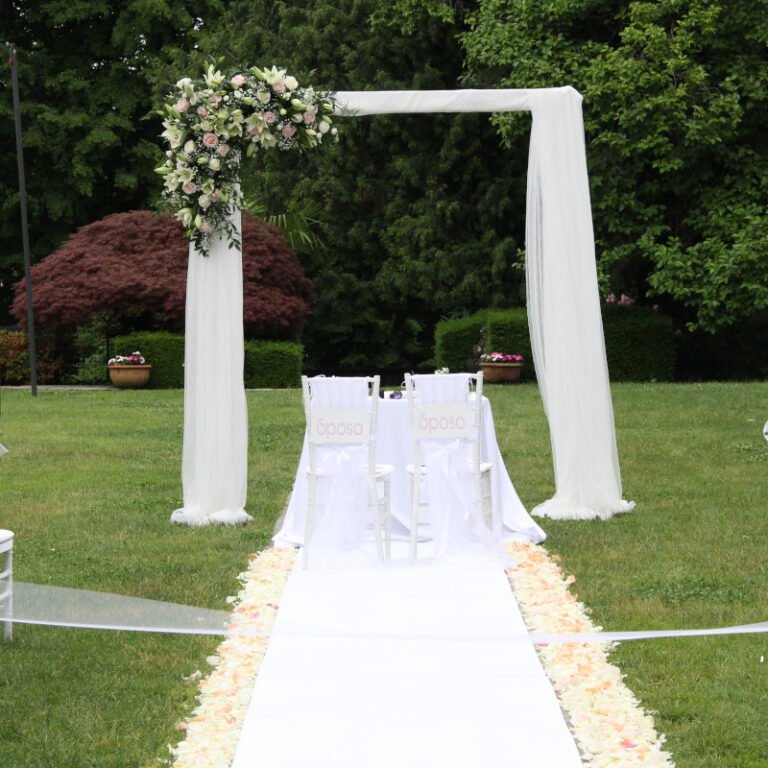 Arredo in alluminio per cerimonia e matrimonio a forma di arco decorato con tulle bianco e fiori ad incorniciare le sedie degli sposi su un prato